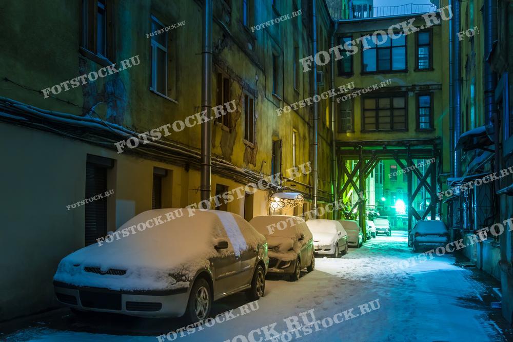 На фото изображен зимний дворик в Санкт-Петербурге. Видны заснеженные автомобили и старые здания, освещенные уличными фонарями. Это фото подойдет для использования в туристических материалах, путеводителях и статьях о зимнем Санкт-Петербурге.